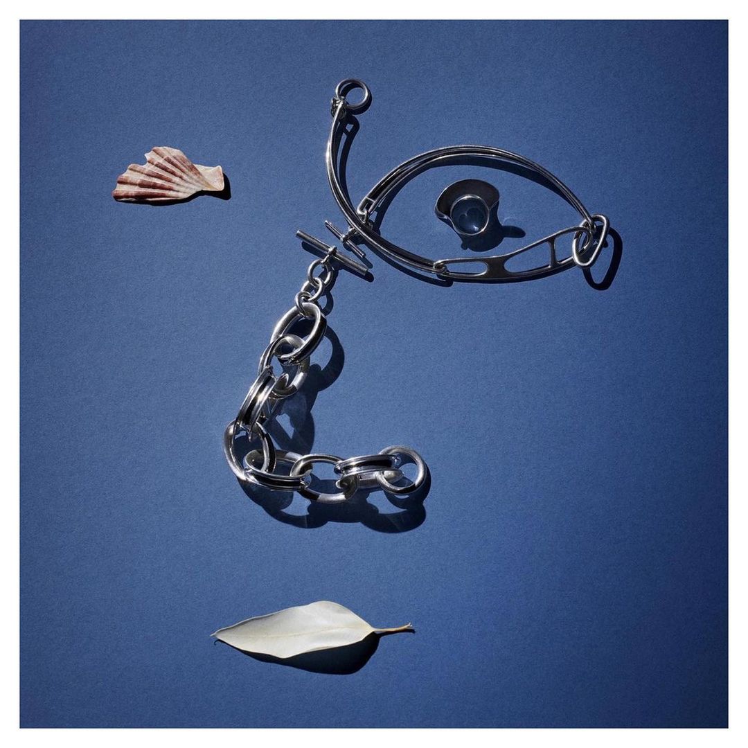 Overhead shot of silver chain necklace, ring, leaf, and seashell arranged on a blue background, illuminated by a strong light source.