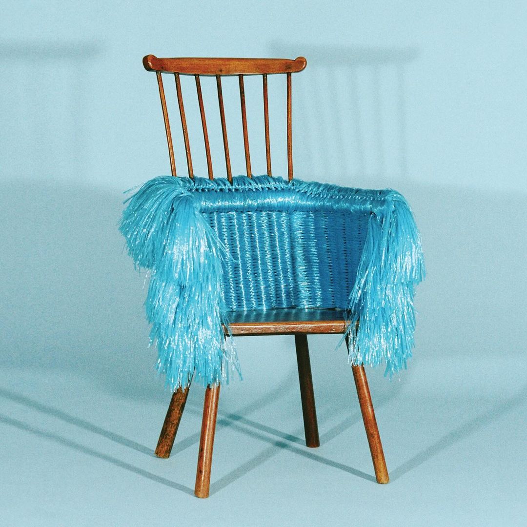 A wooden chair with a blue woven seat and blue fringe is set against a gradient blue background. The chair has a wood frame and legs.