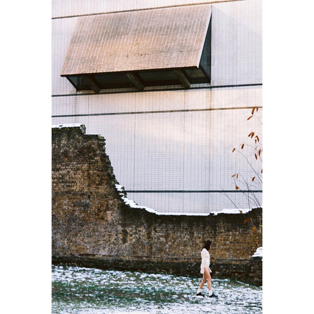 Woman in a white coat walks by a weathered brick wall and a white building with an interesting window structure in an urban area.