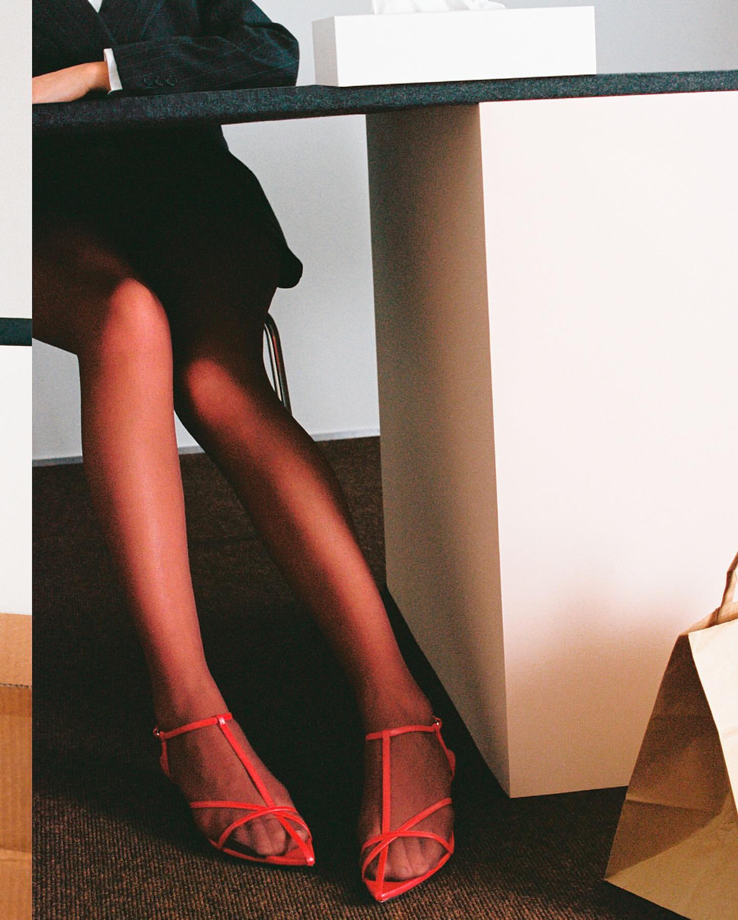 Close-up of a person sitting at a desk wearing red strappy flat shoes and a dark skirt.