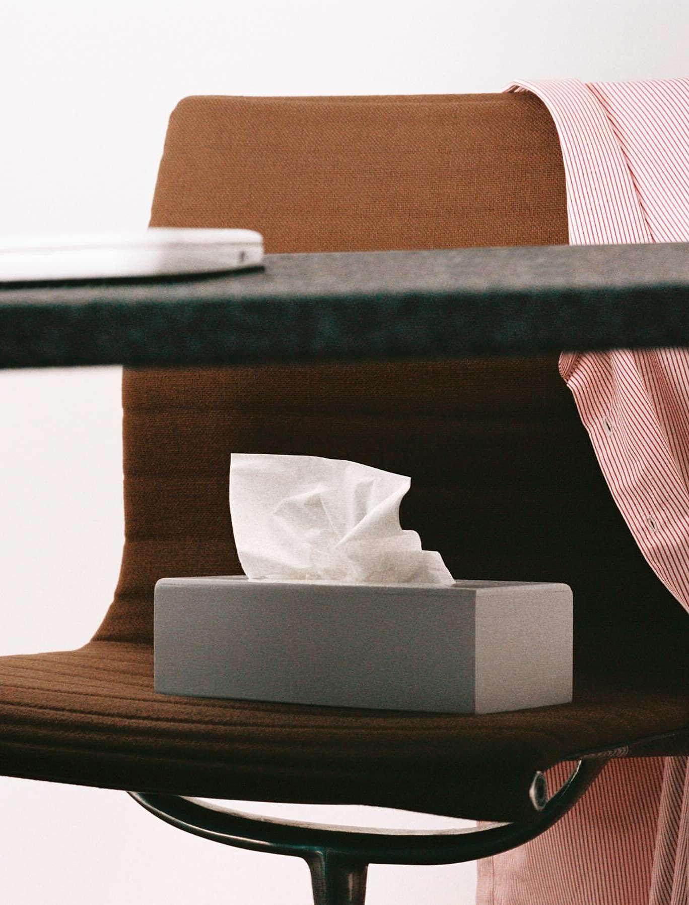 Close-up of a tissue box on a brown chair near a desk with a laptop and a red and white striped shirt. Interior setting.