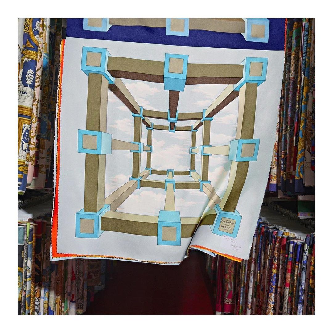 Square silk scarf with an optical illusion design featuring a geometric cube structure against a sky background displayed among fabrics.