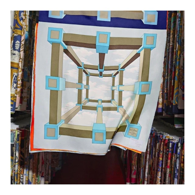 Square silk scarf with an optical illusion design featuring a geometric cube structure against a sky background displayed among fabrics.
