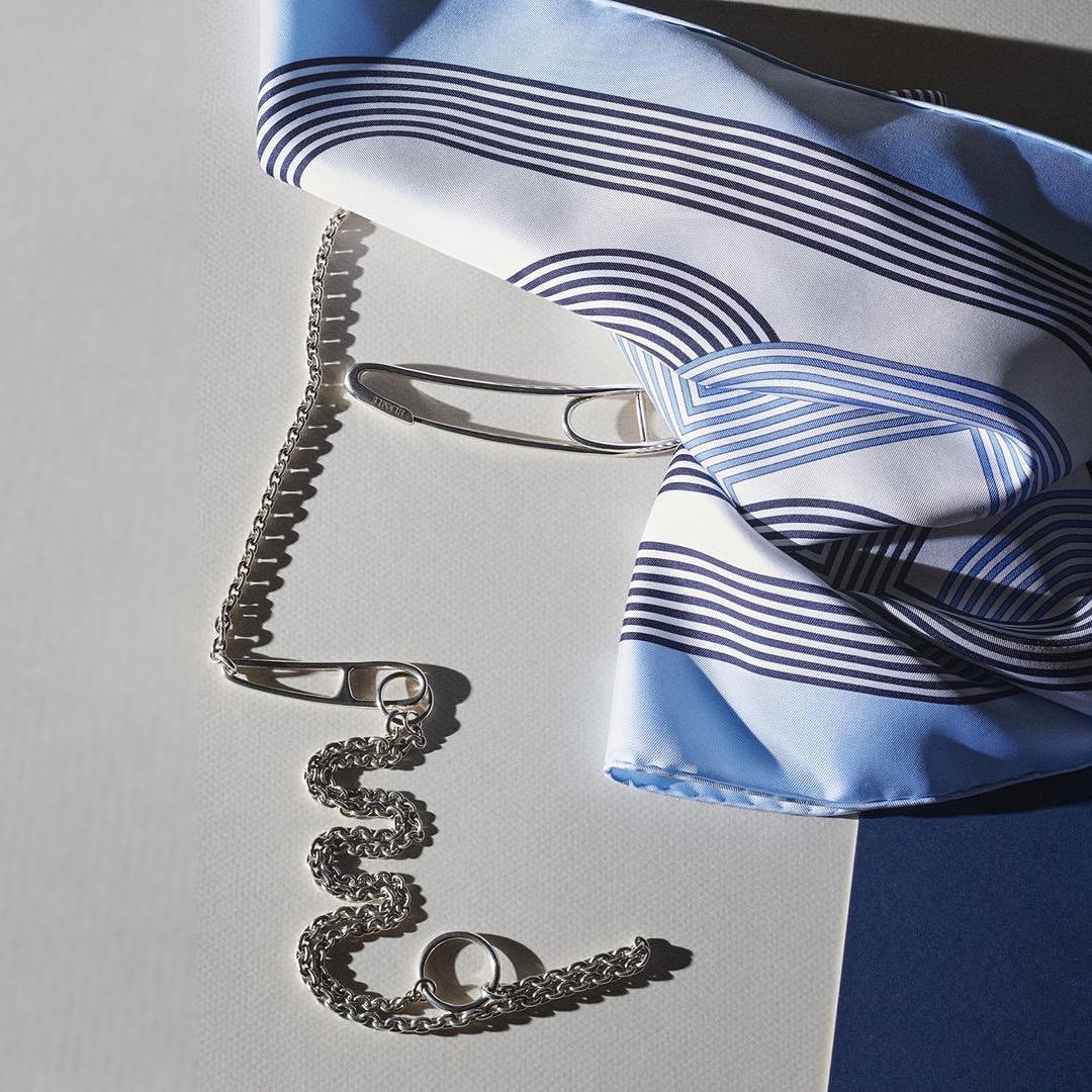 Silk scarf with a blue and white pattern arranged with a silver chain and a safety pin on a gradient white and blue surface.