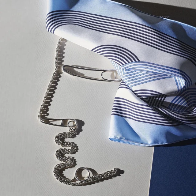 Silk scarf with a blue and white pattern arranged with a silver chain and a safety pin on a gradient white and blue surface.