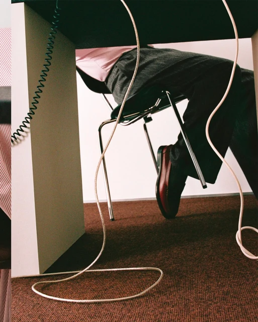 Man sits under a desk with visible legs, feet in brown shoes, grey pants, and light pink shirt, surrounded by cords and brown carpet.