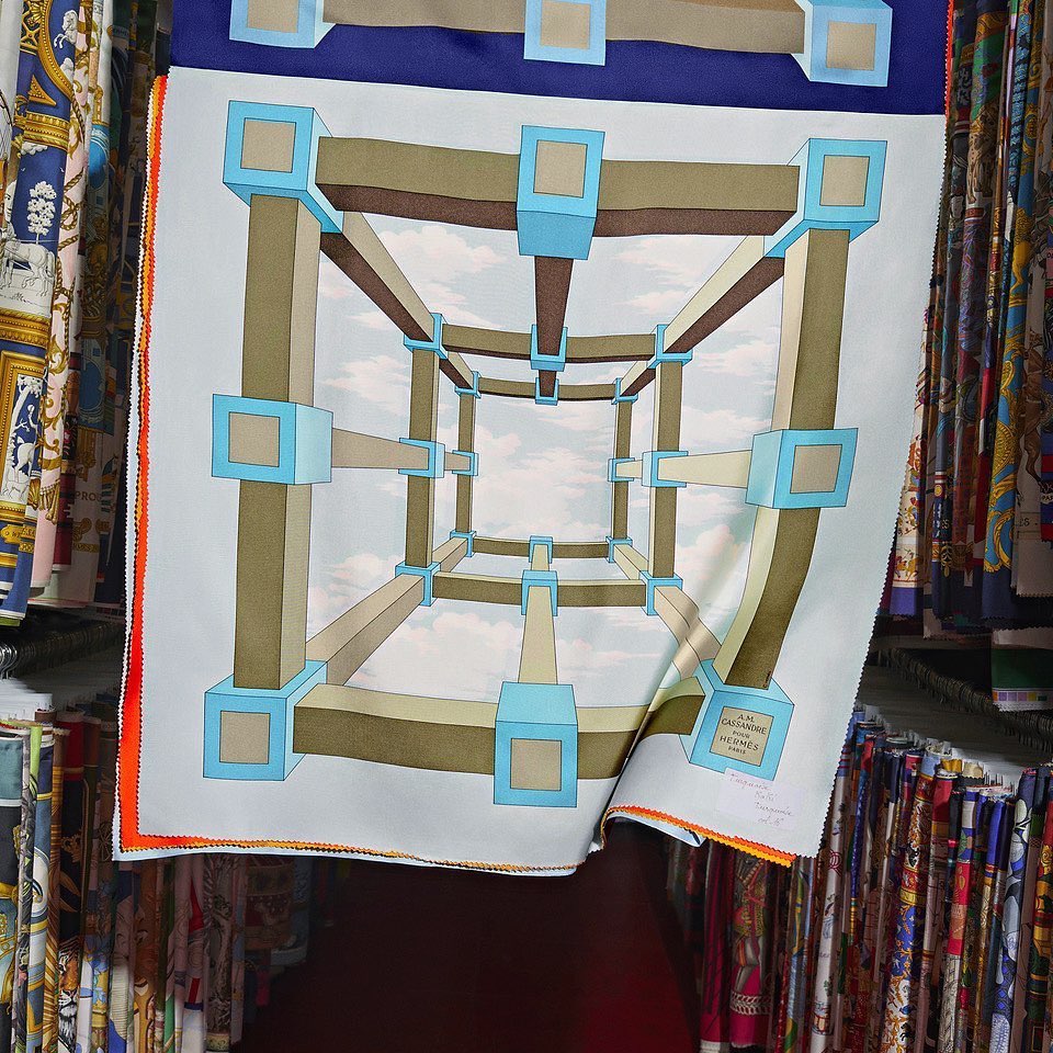 Scarf with a geometric cube design in blue, brown, and white, presented indoors amongst other fabrics. The image has a shallow depth of field.