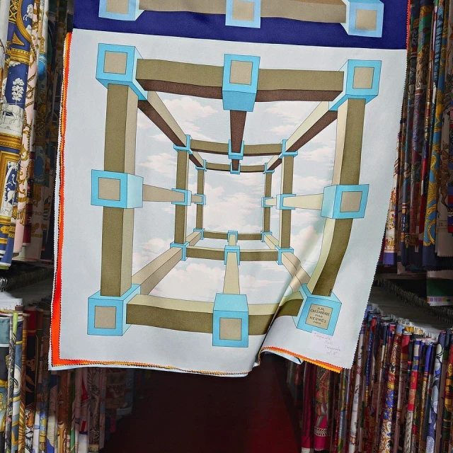 Scarf with a geometric cube design in blue, brown, and white, presented indoors amongst other fabrics. The image has a shallow depth of field.
