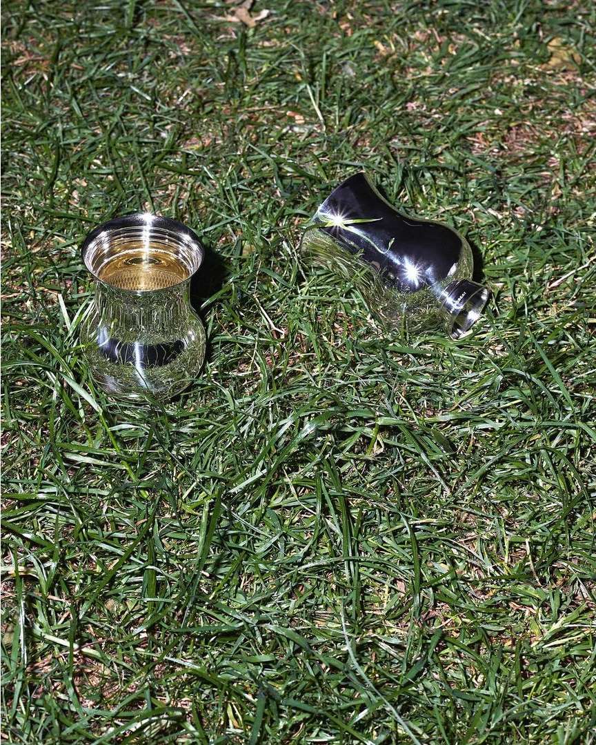Two metallic vases, one upright and one tipped over, sit on bright green grass, reflecting sunlight and their surroundings.