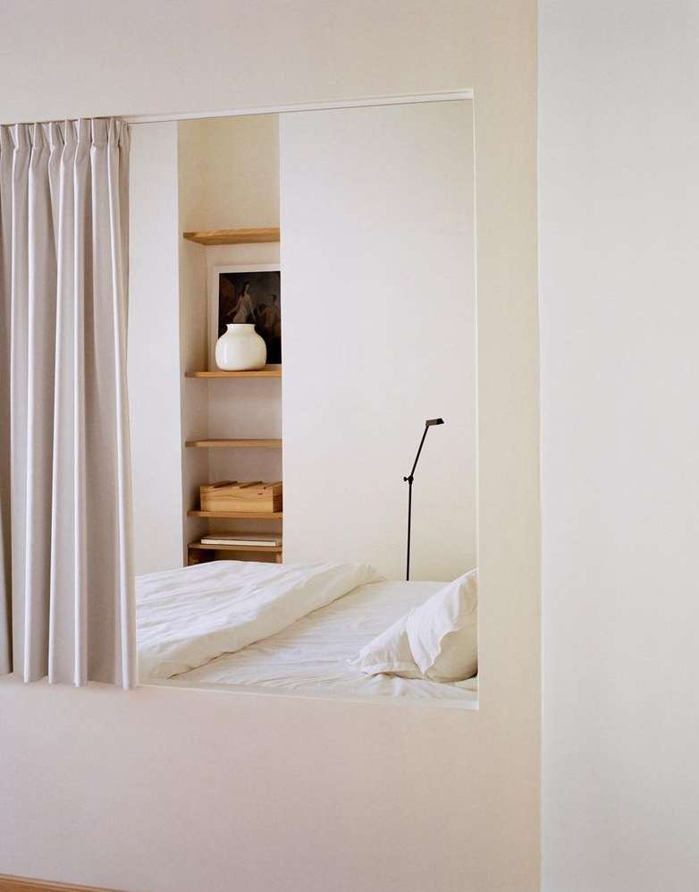 Minimalist bedroom interior featuring a bed, curtains, built-in shelves, decorative vase, and lamp, set in a bright, naturally lit space.
