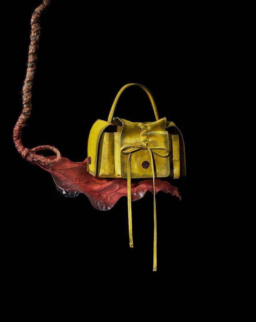 Yellow leather purse with top handle and drawstring closure sits on a red leaf against a dark background in a studio product shot.
