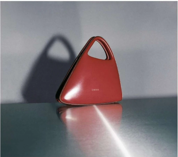 Minimalist product shot: A rust-colored handbag with a triangular shape and handle is displayed on a reflective surface against a gray wall.