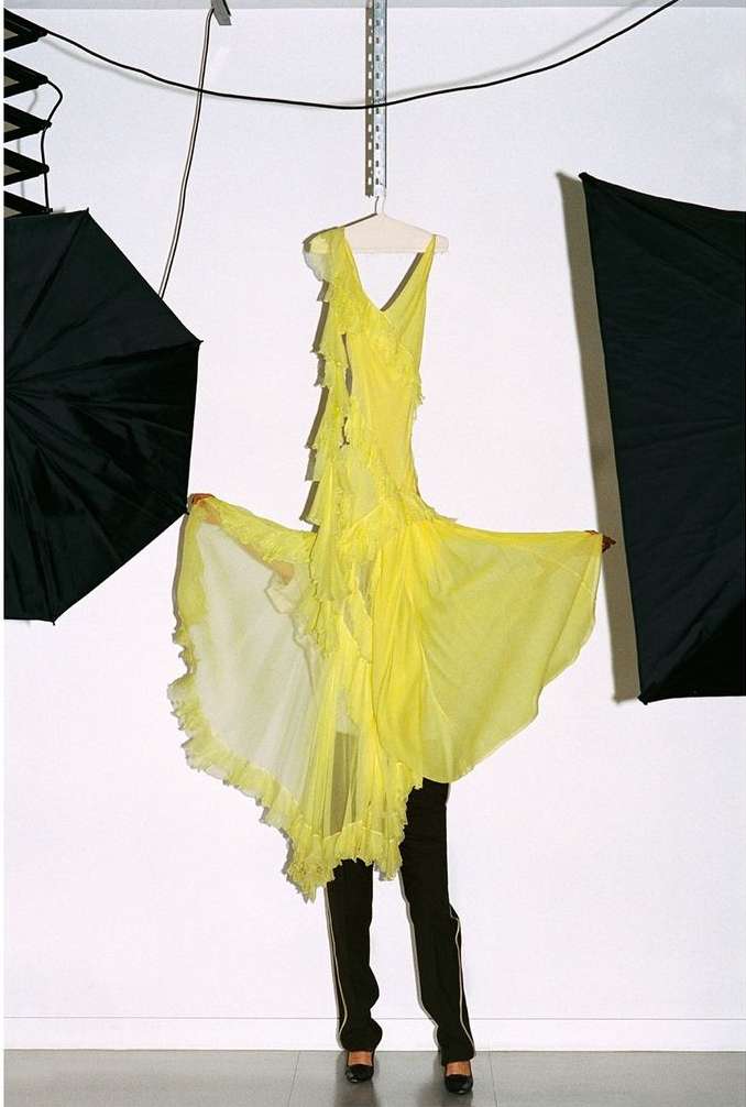 Bright yellow ruffled dress on a hanger, partially obscured by a person in dark pants and heels, in a photo studio with lighting equipment.