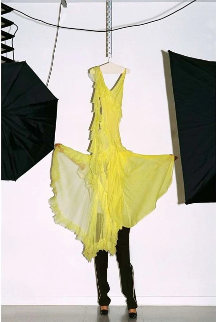 Bright yellow ruffled dress on a hanger, partially obscured by a person in dark pants and heels, in a photo studio with lighting equipment.