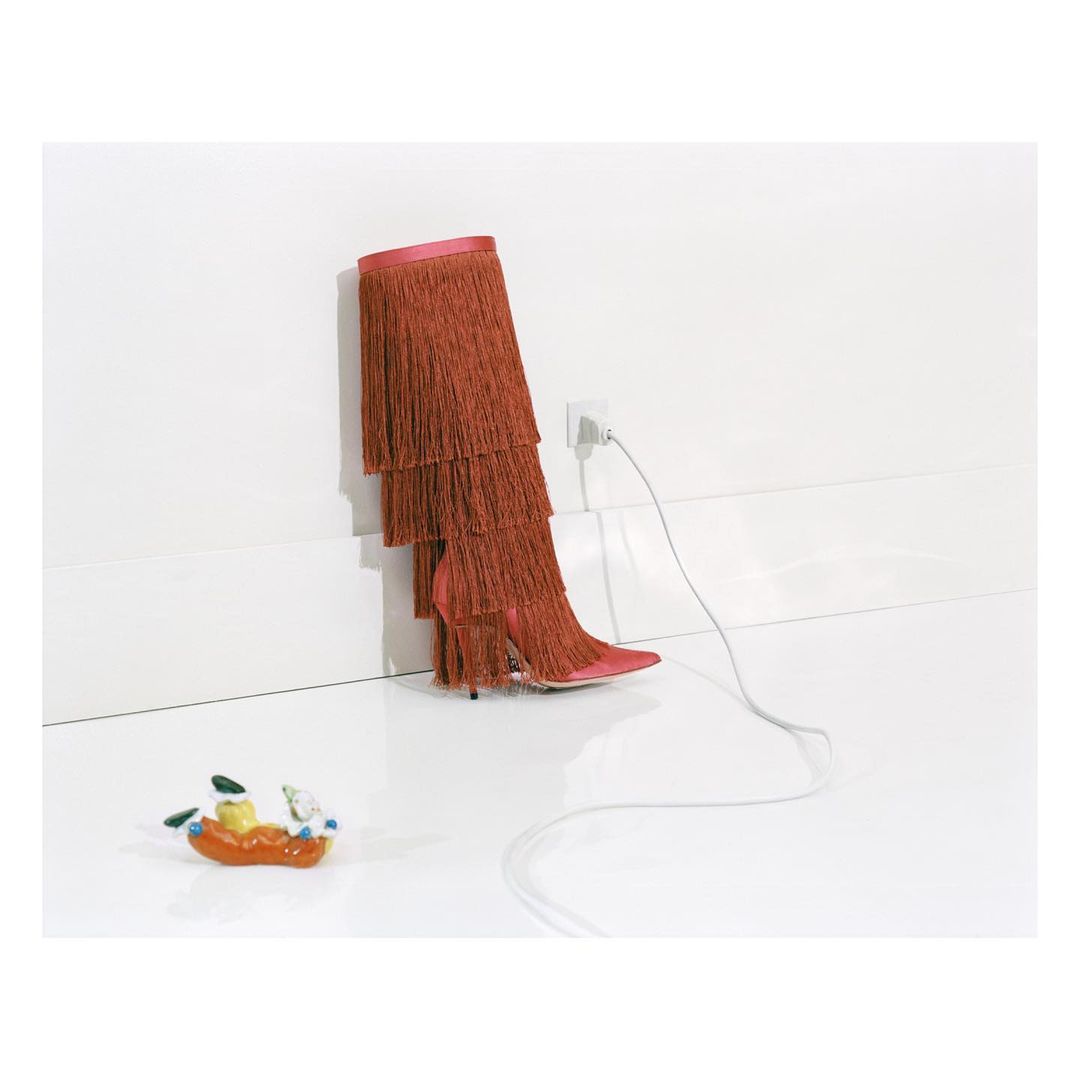 High-heeled boot with orange fringe stands plugged into a wall socket next to a ceramic figurine on a white floor against a white wall.