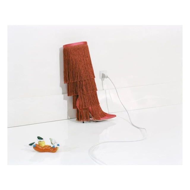 High-heeled boot with orange fringe stands plugged into a wall socket next to a ceramic figurine on a white floor against a white wall.