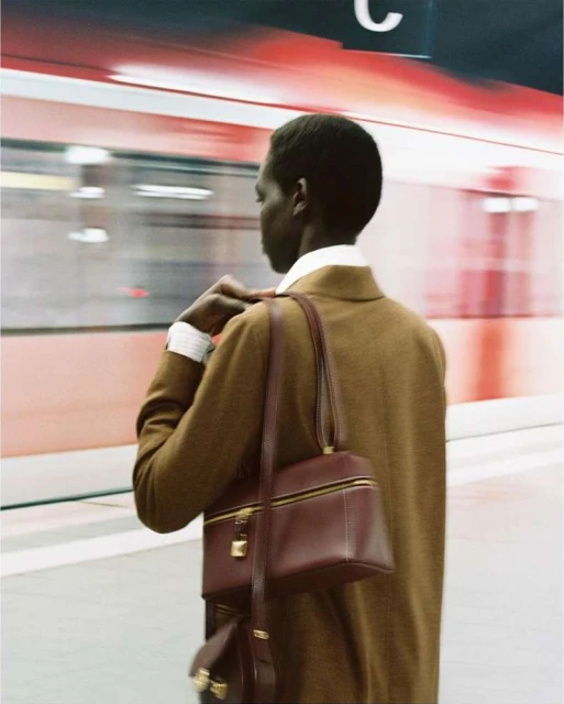 A person stands on a subway platform with a red train blurred in motion behind them, wearing a brown jacket and carrying a brown bag.