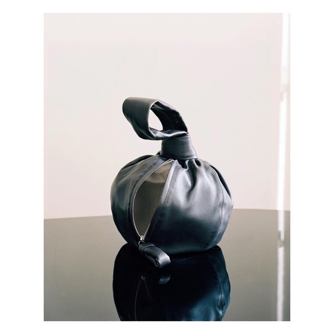 Elegant black leather dumpling bag with knotted handle and zipper closure on reflective black surface against a white background.