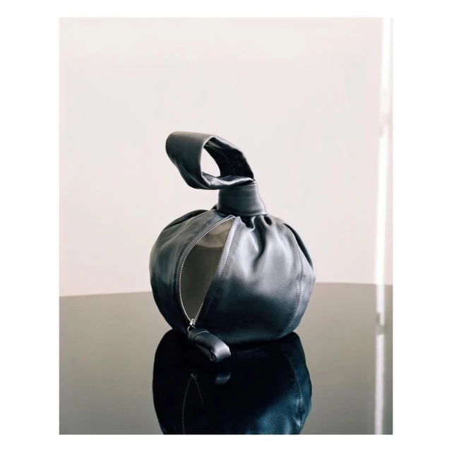 Elegant black leather dumpling bag with knotted handle and zipper closure on reflective black surface against a white background.