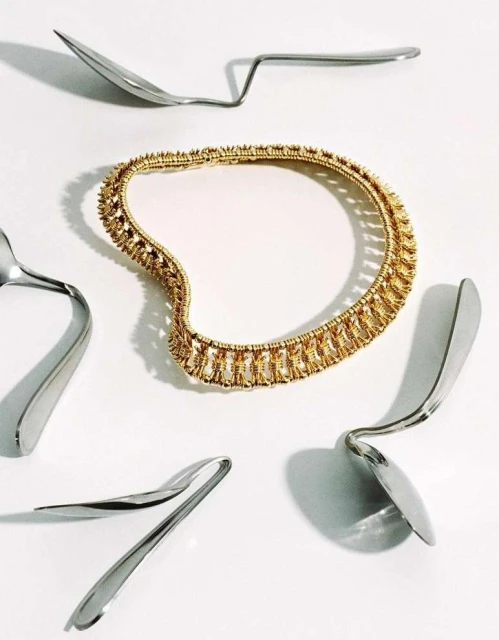A gold necklace is presented surrounded by the reflective silver surfaces of spoons on a plain white backdrop in a studio setting.