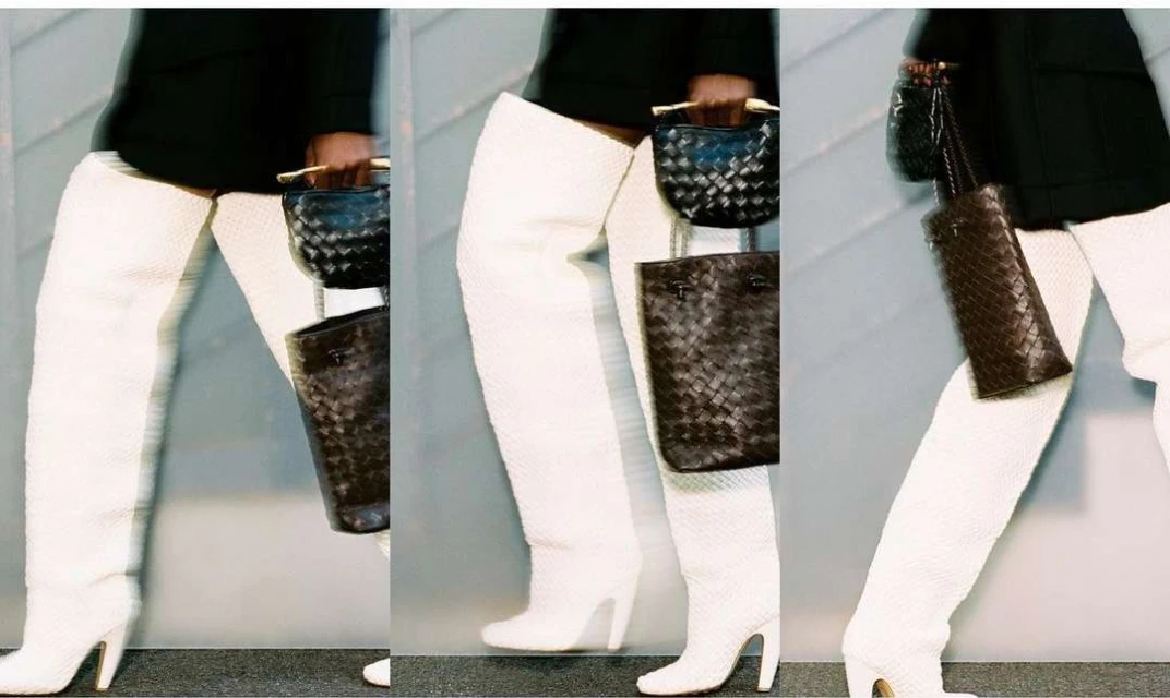 Fashion shot featuring a person with a black jacket holding small woven handbags, wearing white textured over-the-knee high-heel boots, indoors.