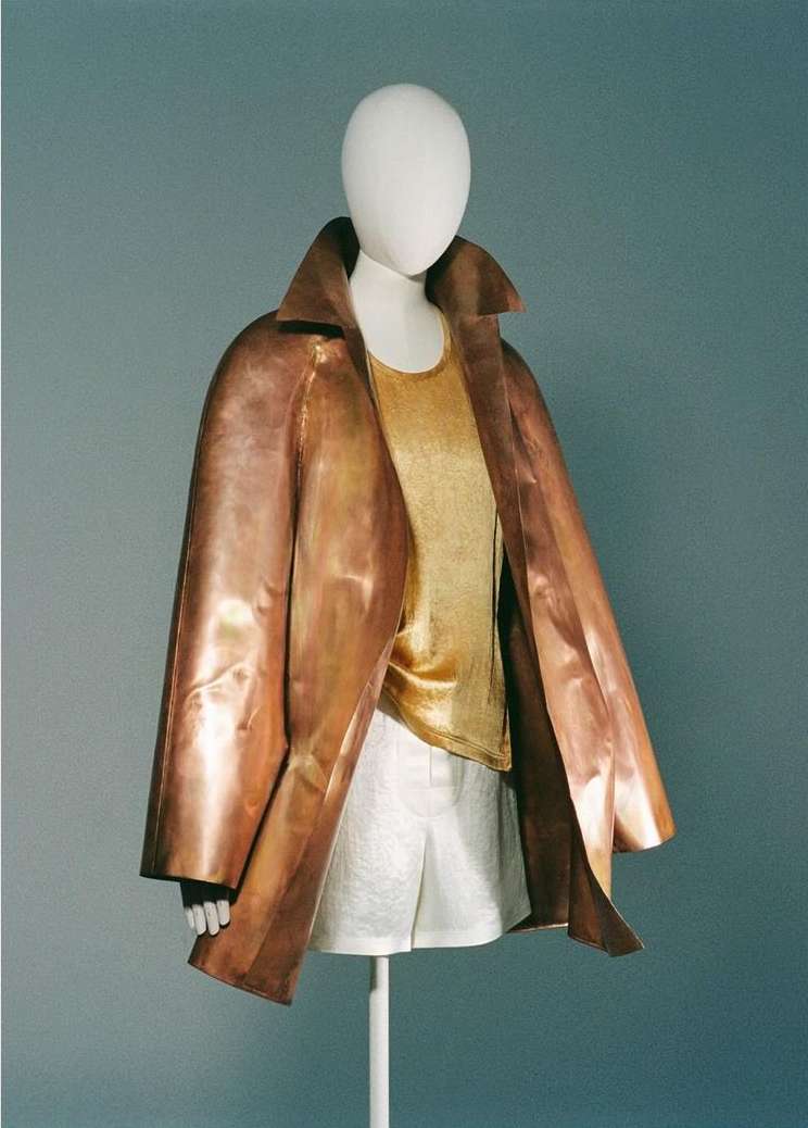 Mannequin displays copper coat and gold top with white skirt on pale blue. The display is minimalist and fashion-forward.