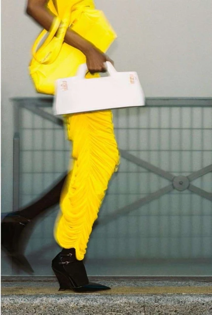 Fashionable woman walks wearing yellow dress and black heels, carrying a white purse against gray wall. Motion blur creates a sense of movement.