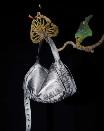 Studio shot of a metallic silver handbag with the word ACNE hanging from a decorative branch against a solid black backdrop.