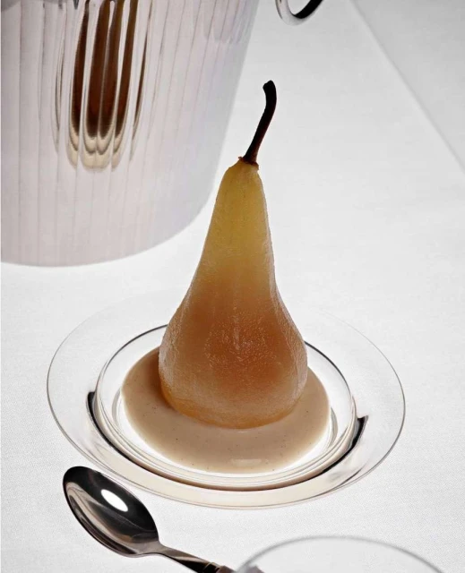 Poached pear dessert on a clear glass plate with sauce, a silver spoon, and a bucket, set against a crisp white background.