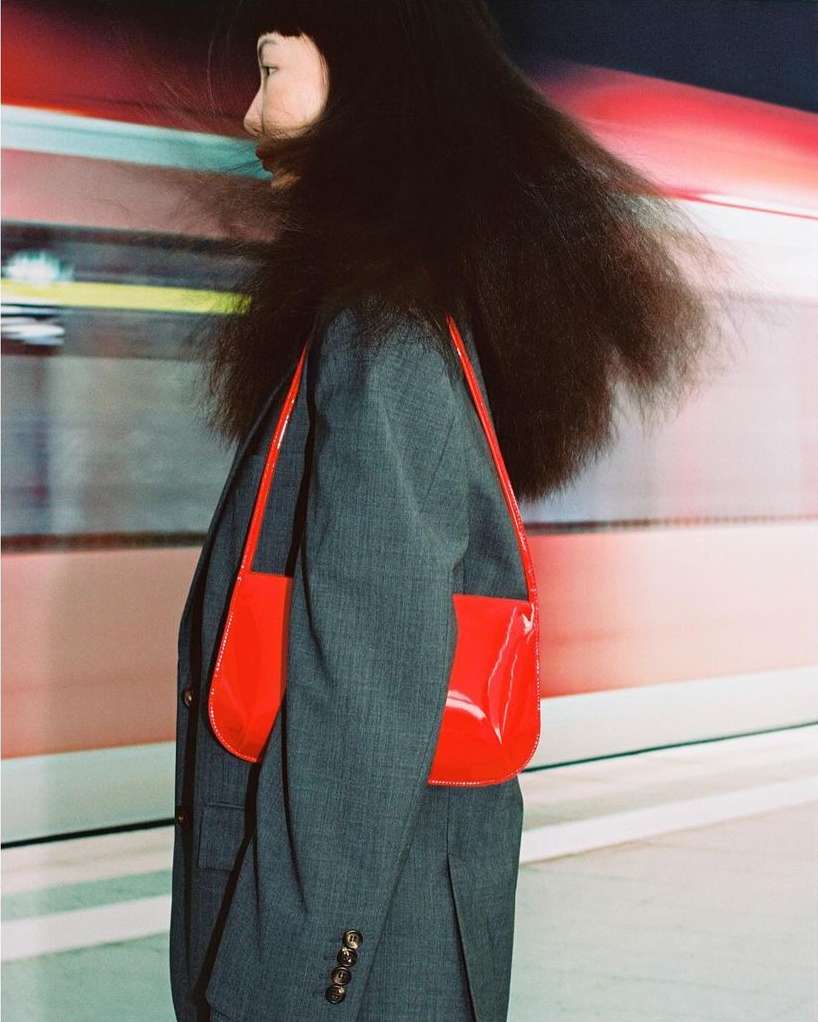Woman with long dark hair and grey suit with a bright red patent leather purse. Motion blur in the background.
