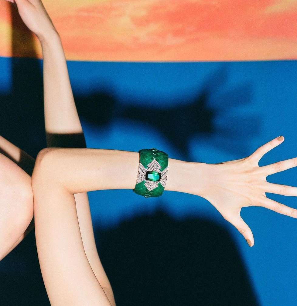 Woman's arms and hands shown with a green and gemstone cuff bracelet against a color-blocked orange and blue background.