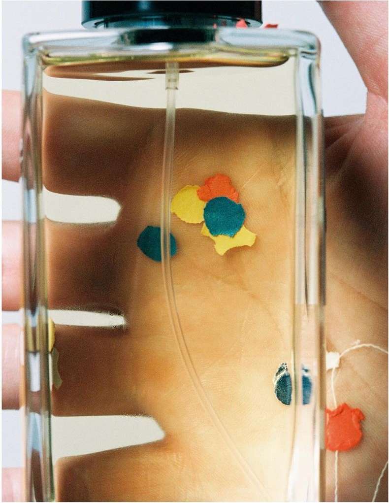 Close-up of a person's hand holding a clear glass perfume bottle with a black cap, featuring colorful abstract spots inside the bottle.