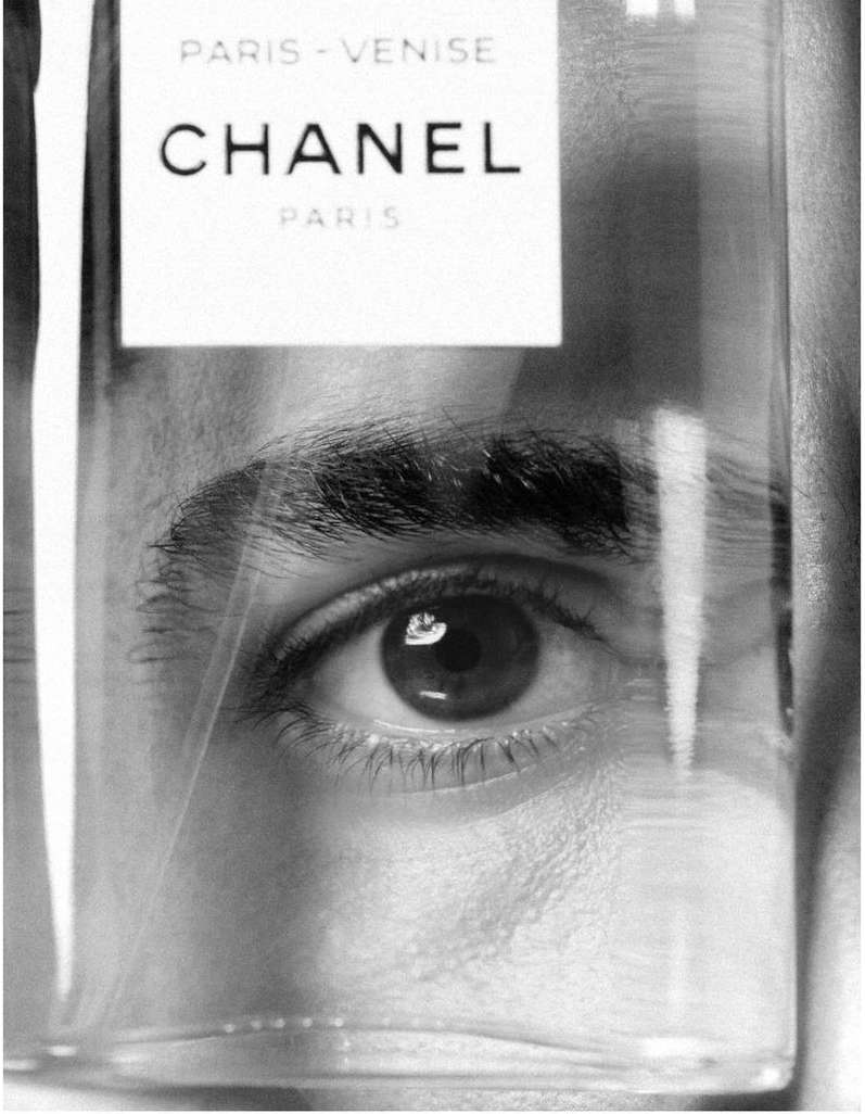 Monochrome close-up of a person eye viewed through a transparent Chanel Paris Venise perfume bottle.