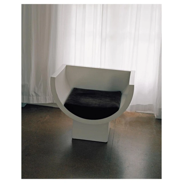 Minimalist interior featuring a sculptural white chair with a dark grey cushion against white curtains and a polished grey floor.