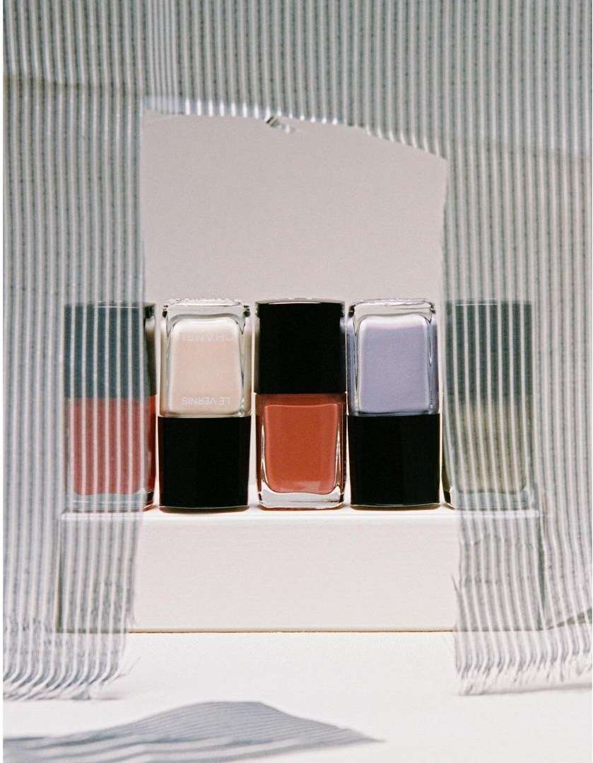 Three bottles of nail polish in pink, coral, and lavender, arranged on a white platform against a striped fabric background.