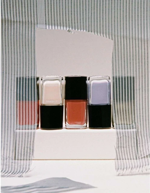 Three bottles of nail polish in pink, coral, and lavender, arranged on a white platform against a striped fabric background.