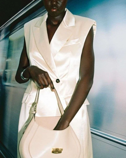 Dark-skinned woman in a sleeveless white blazer dress holds a cream-colored purse in an indoor setting with diffused lighting.