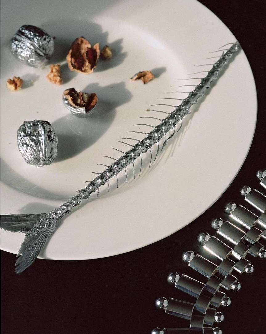 Still life shot of a metal fish skeleton on a white plate with whole and cracked walnuts on a dark surface.