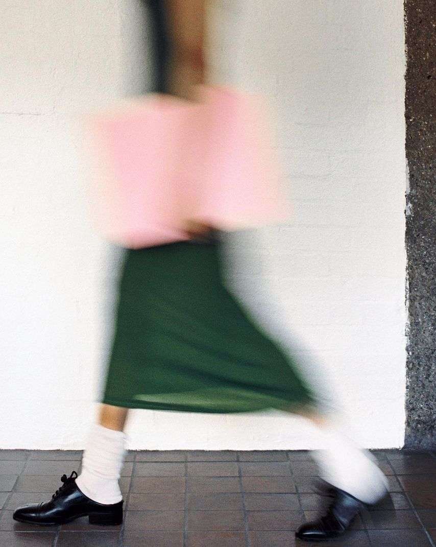 Motion blurred fashion photograph of a person walking wearing a green skirt, white socks, and black shoes, carrying a pink bag.