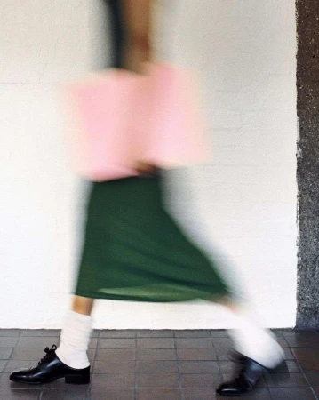 Motion blurred fashion photograph of a person walking wearing a green skirt, white socks, and black shoes, carrying a pink bag.