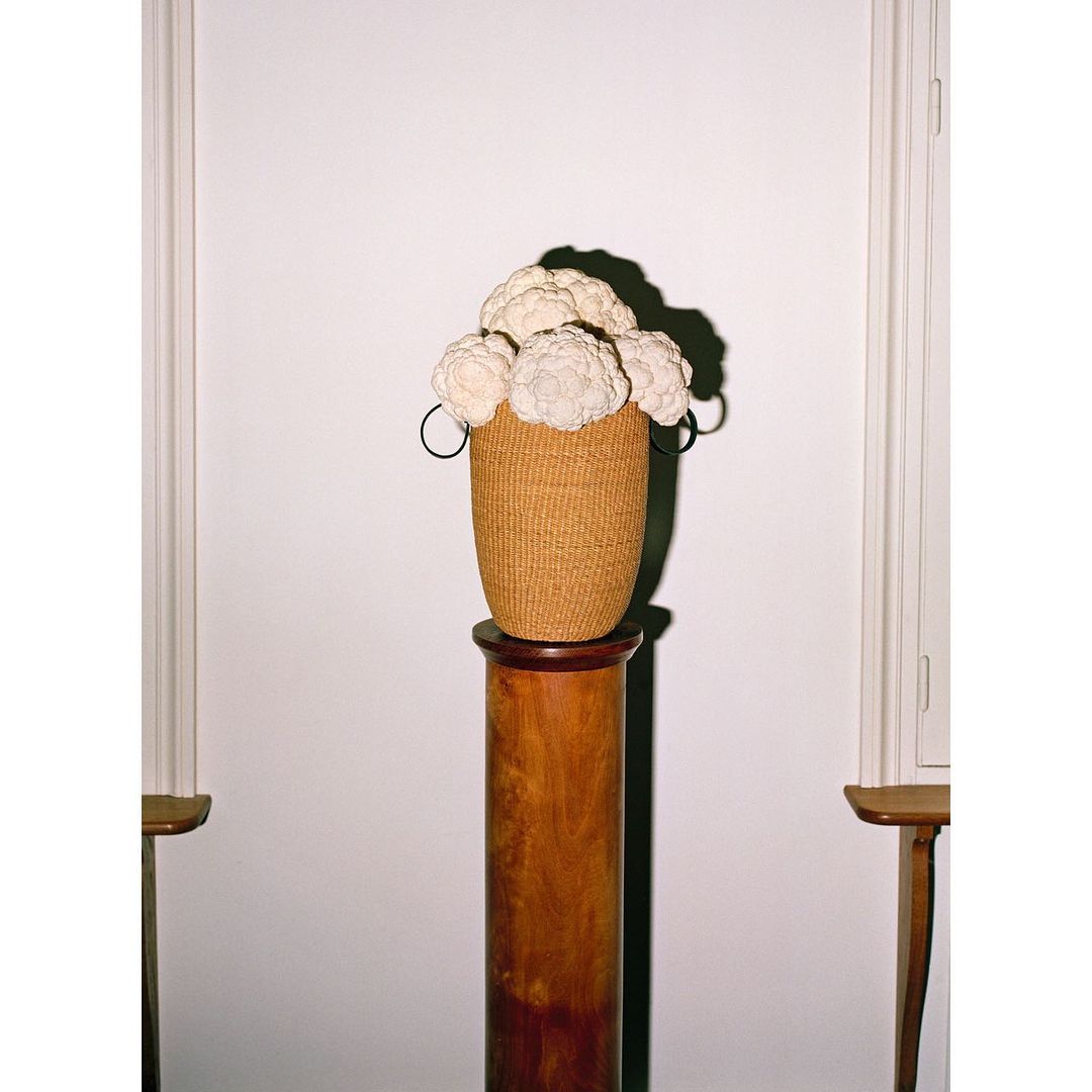 Tan woven basket filled with white textile flowers placed on a wood pedestal against a white wall with partial window and door frames.