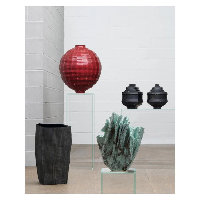 Still life of various sculptural vases in red, black, and green displayed on clear acrylic pedestals in a minimalist white gallery setting.
