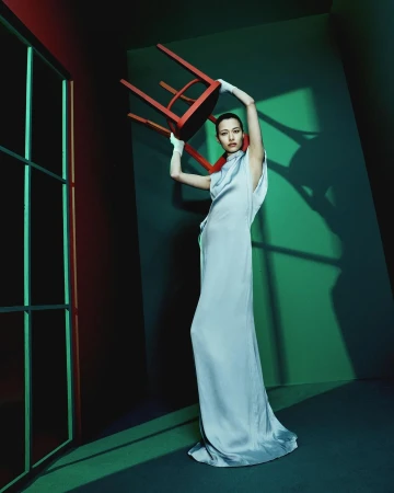 Model in a silver satin dress holding a red wooden chair overhead in a styled studio with dramatic lighting.