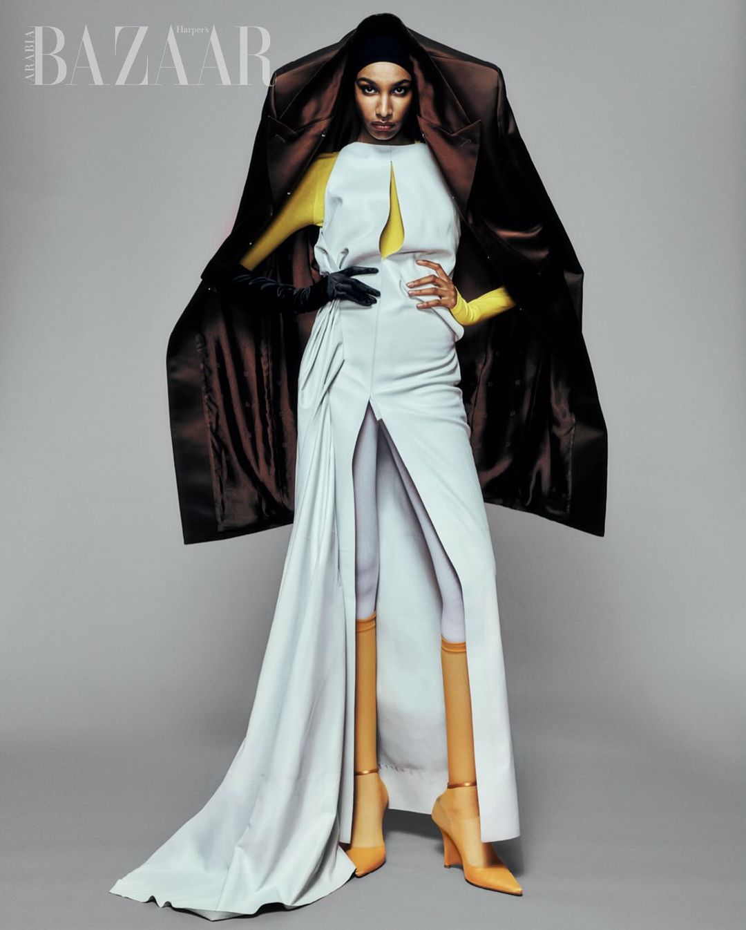 Fashion editorial shot of a woman in a long white dress and brown coat against a grey background, showcasing high fashion design.