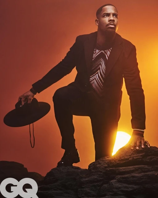 Portrait of a Black man in a suit kneeling on rocks, holding a fedora hat against an orange sunset backdrop and the GQ logo.