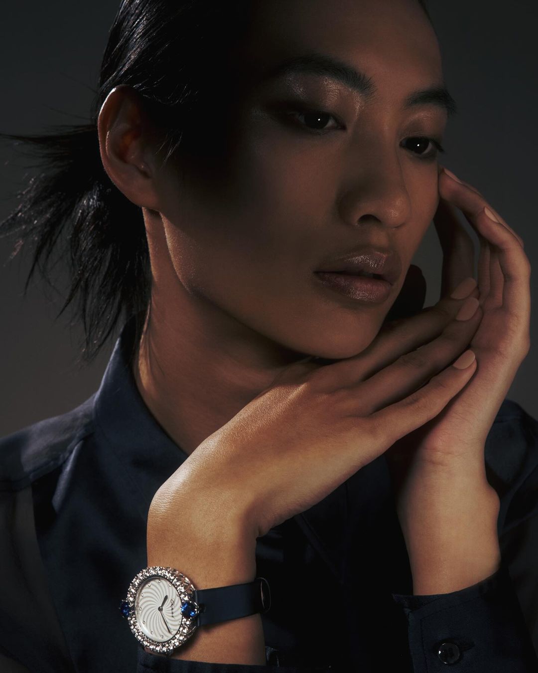 Portrait of a woman with dark hair, wearing a dark blouse and a wristwatch with a decorative bezel and band in a studio setting.