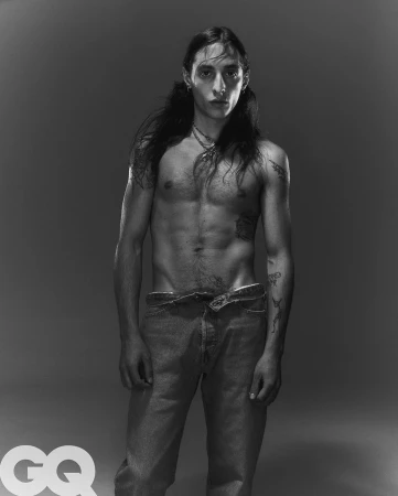 Portrait of a shirtless man with long dark hair wearing jeans against a neutral studio background by Mattia Guolo
