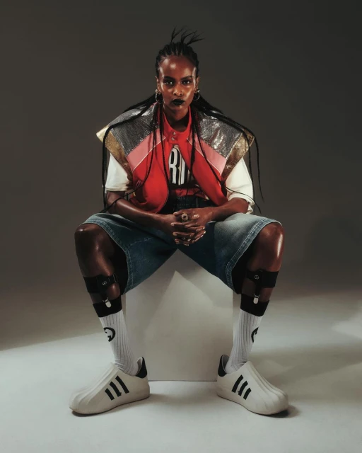 Stylish woman in modern fashion sits indoors: bold makeup, braids, oversized denim shorts, socks with graphics, and Adidas sneakers.