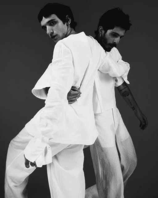 Two men in white shirts and pants stand with their backs partially to each other, posing in a black and white studio portrait.