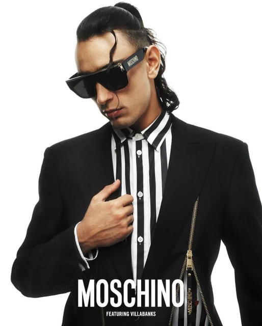 Adult male poses indoors wearing a striped shirt, black jacket, and sunglasses, with a Moschino logo in white against a white background.